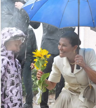 Crown Princess Victoria of Sweden mixes summer style with autumn weather as she holds a bright blue umbrella to protect her cream suit from the rain.
