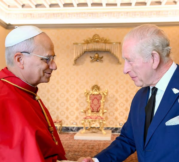 King Charles, in a blue suit, shakes hands with Pope Leo who is wearing a red cape.