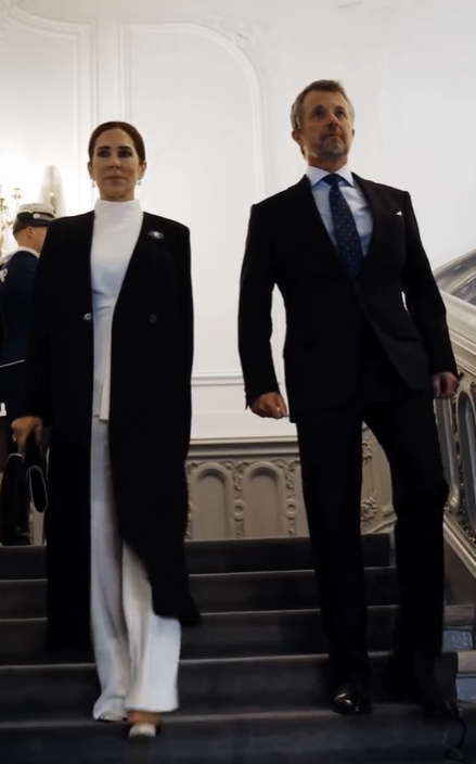 Queen Mary of Denmark walks down palace stairs in a white trouser suit and black coat