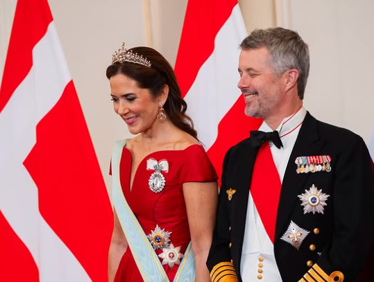 Queen Mary of Denmark wears a red dress with diamond tiara and stands n front of a Danish flag