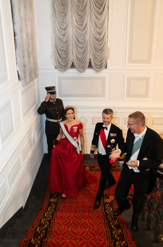 Queen Mary of Denmark, wearing a red ballgown and diamond tiara, walks up a sweeping staircase covered in red carpet