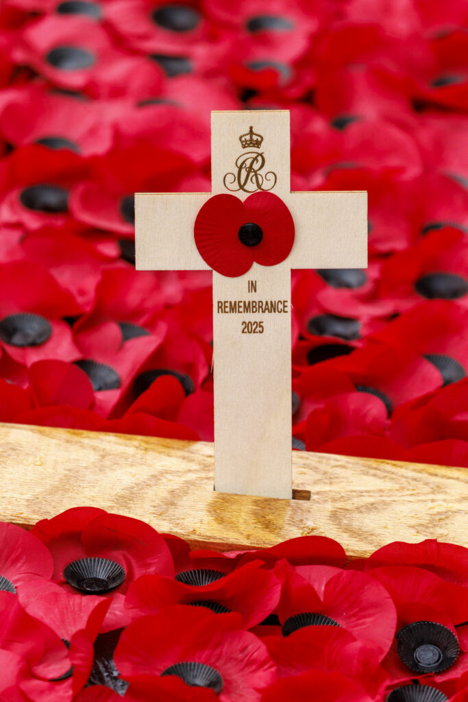 A close up of the wooden cross with a single red poppy and the cypher of Queen Camilla, planted at the Field of Remembrance