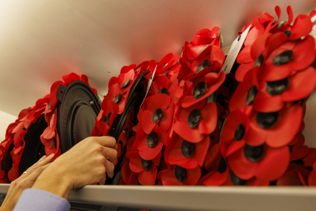 A red poppy wreath is placed on an overhead shelf in a train carriage alongside other brightly coloured tributes