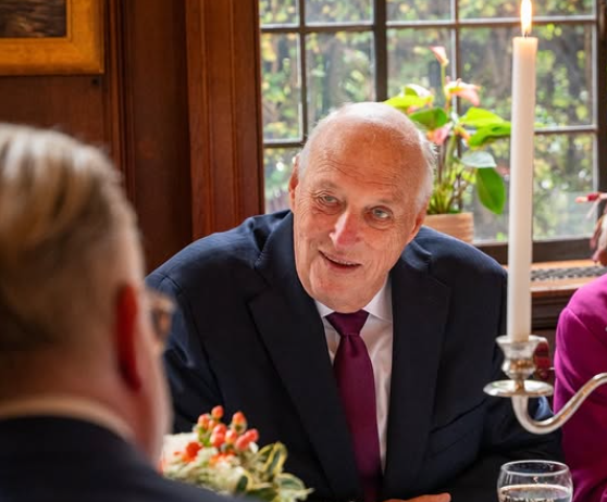 King Harald of Norway sits at a table and talks to a man opposite him. A candle in a silver candlestick is seen in the foreground.