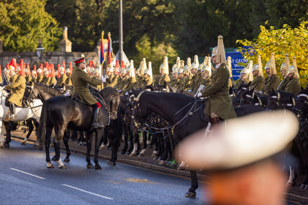 Troops in khaki uniform sit on horses who are standing on a road