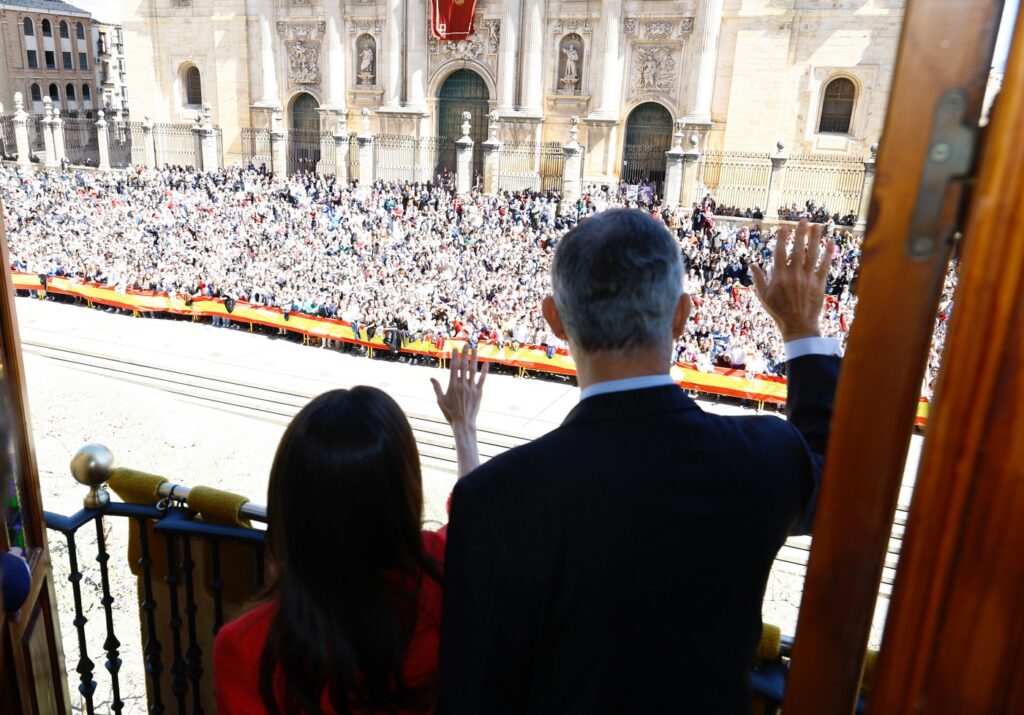 King Felipe and Queen Letizia are seen in shadow as they wave from a balcony in Jaen