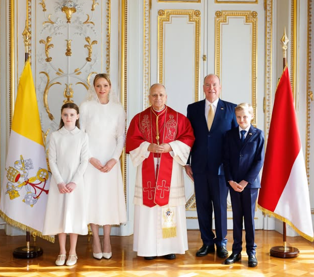 Pope Leo XIV stands in front of the flag of Monaco and of the Vatican on a meeting with Monaco's royals