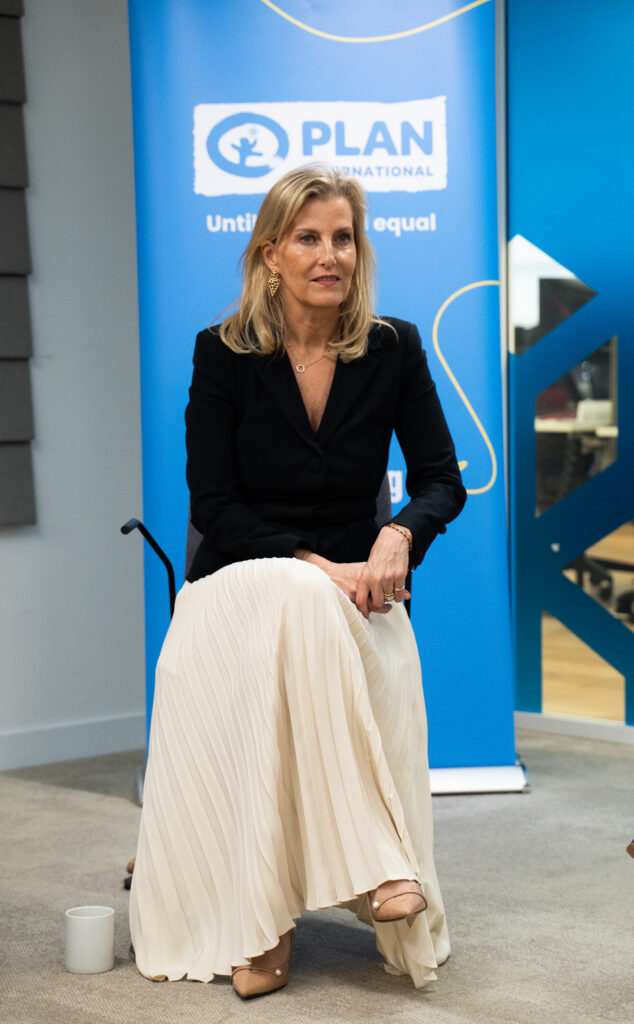 The Duchess of Edinburgh, in a black jacket and white skirt, sits in front of a Plan International banner