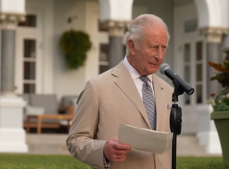 King Charles wears a pale jacket and tie as he stands in a beautifully manicured garden in Bermuda