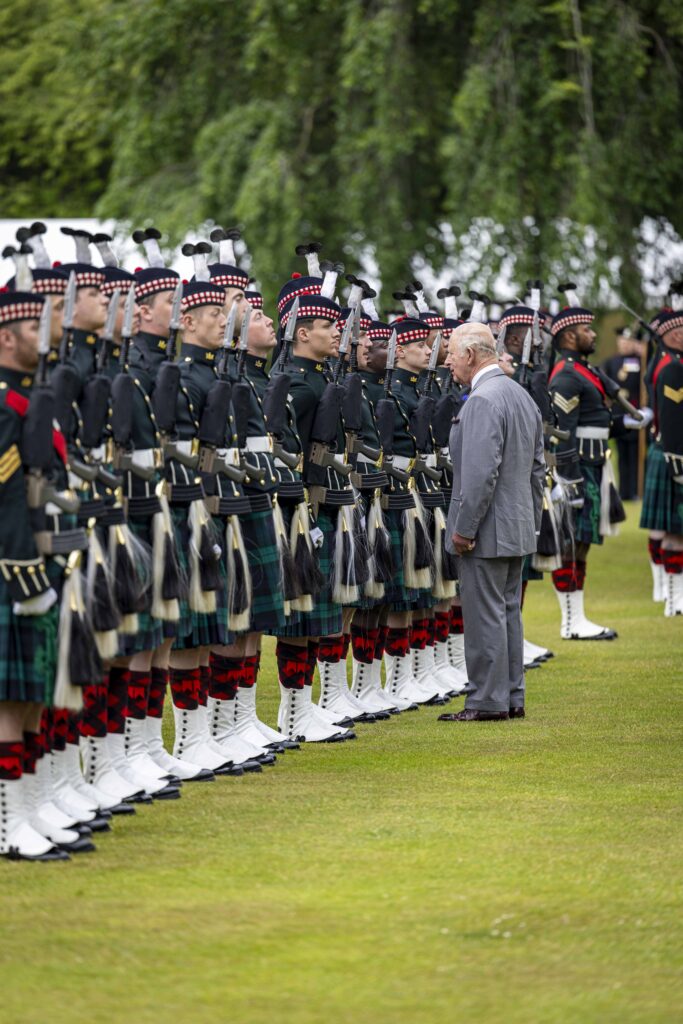 King Charles, in a grey suit, speaks to soldiers wearing kilts